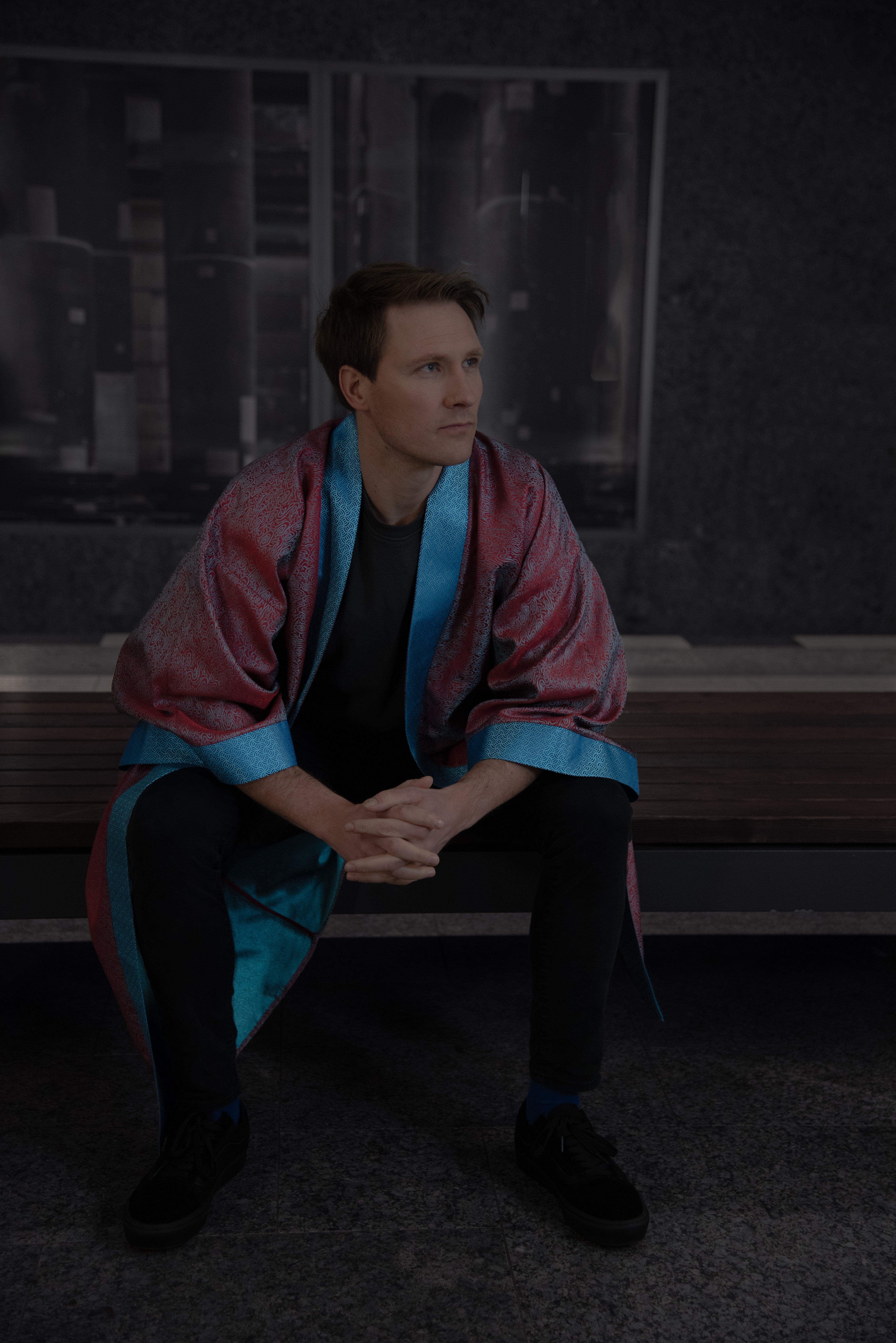 Model wearing a Burgundy coloured Kimono with botanical details (front view, model sitting), showcasing burgundy red fabric, turquoise blue collar, arm bands and reflections of the fabric on the inside.