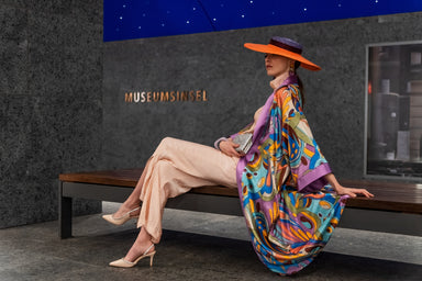 A model sitting on a bench, wearing an orange and lilac coloured hat and nude onesie. The Kimono is visible from the side and drapes down softly onto the bench. Delicate florals, bursting in a rainbow of colors, adorn the softest mulberry silk. A silky, lilac coloured collar and arm bands can be seen.